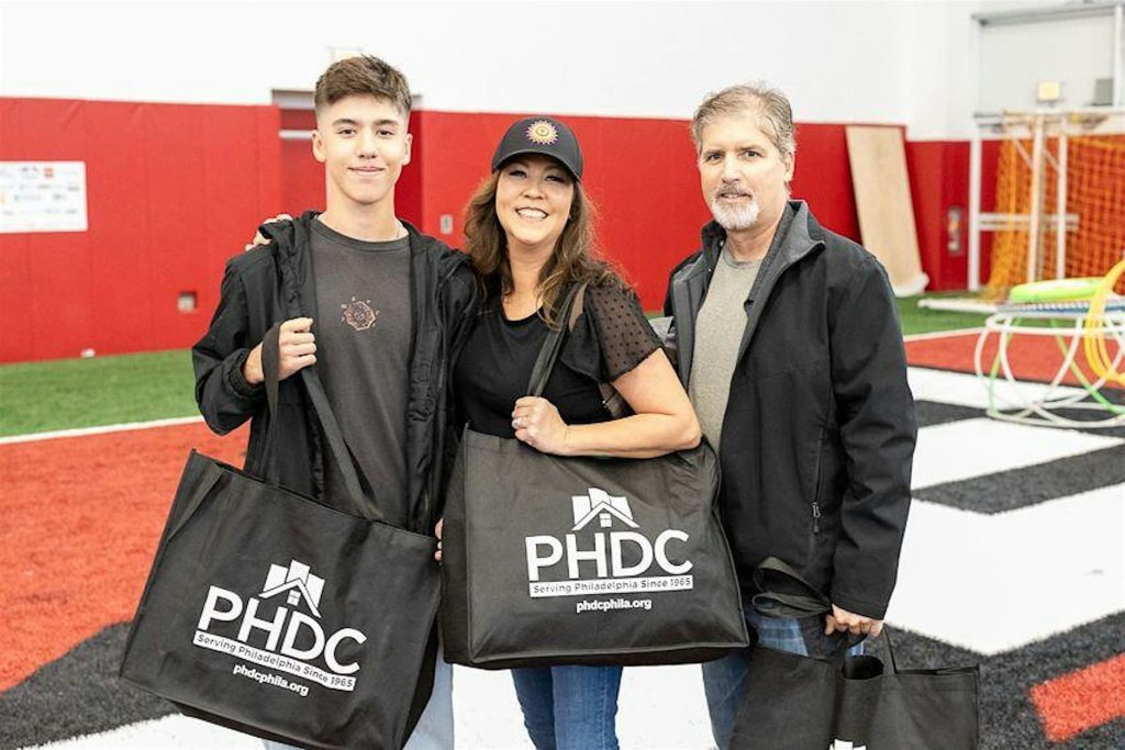Three people holding black tote bags labeled 'PHDC' inside a brightly lit indoor sports facility with artificial turf.